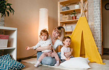 a happy mother with two twin girls plays at home on the floor sitting near a yellow teepee. joint games of mothers and daughters.の写真素材