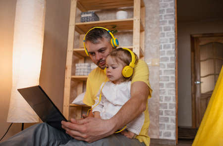a happy father and his little daughter are sitting on the floor with yellow headphones and looking at a laptop. the concept of family happinessの写真素材