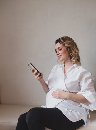 A pregnant woman keeps and uses her smartphone at home. Close-up. A young pregnant woman holds her stomach and looks at her mobile smartphone. Pregnancy.の写真素材