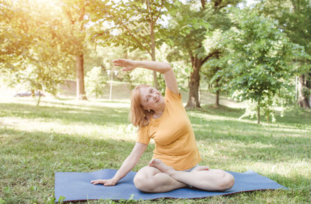 A happy mature woman in a yellow T-shirt is doing yoga, sitting in the park in the lotus position,...