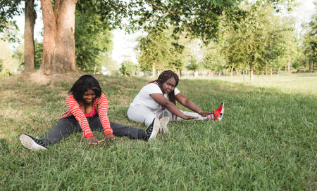 Two African-American girls are engaged in sports, doing exercises sitting in the park on the grass....