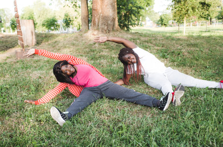 Two African-American girls are engaged in sports, doing exercises sitting in the park on the grass. healthy lifestyleの写真素材
