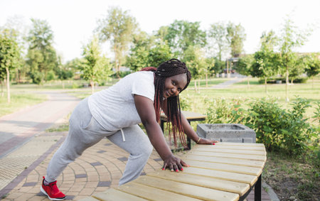 an African-American girl is doing sports warming up near a bench in an outdoor park. healthy...