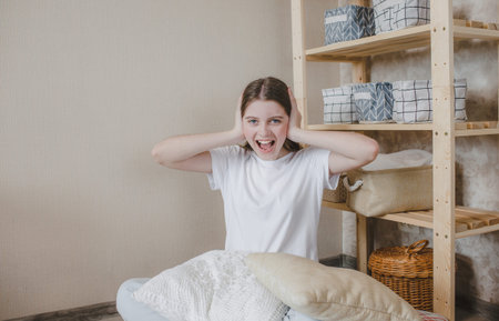 a young girl screams loudly holding her head while sitting on the floor among pillows. anxiety and...
