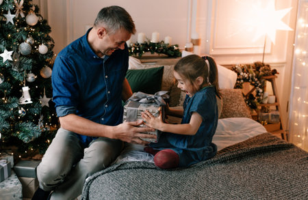 A young father gives a Christmas present to his smiling daughter near a decorated Christmas tree.の写真素材