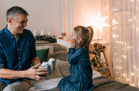 a caring father gives a Christmas gift to his little daughter sitting on the bed near the Christmas tree.の写真素材