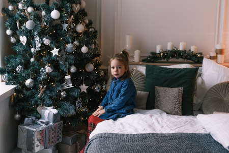 Portrait of a frustrated and little girl sitting on a bed near a Christmas tree.の写真素材