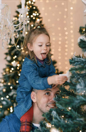 Happy dad and daughter decorate the Christmas tree. A little girl is sitting on Daddy's shoulders, holding a toy on the Christmas tree.の写真素材