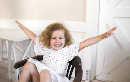 a beautiful little curly-haired girl is sitting on a chair smiling with her arms outstretchedの写真素材