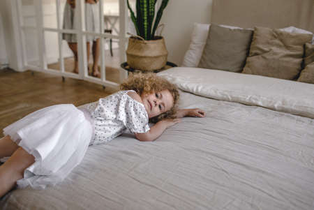 a beautiful little curly-haired girl is lying on a large bed looking awayの写真素材