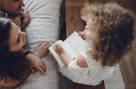 a little girl is reading a book for her parents who are lying on the bedの写真素材
