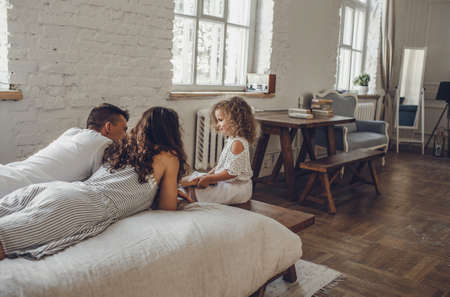 a little curly-haired girl is sitting next to the bed on which dad and mom are lying. family weekendの写真素材