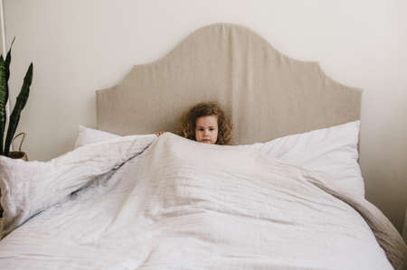 Cute charming curly-haired baby girl lying in bed on a white pillow. The concept of childhood.の写真素材