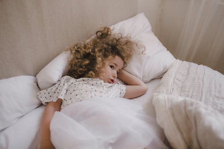 Cute charming curly-haired baby girl lying in bed on a white pillow. The concept of childhood.の写真素材