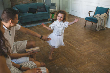 a little girl with curly hair is dancing barefoot on the floor in front of her dad and mom. happy childhoodの写真素材
