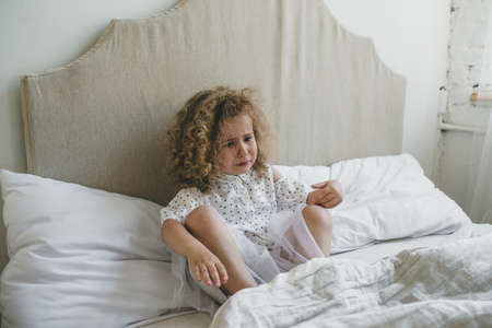 a little girl with beautiful curly hair is crying sitting in bed. upset sad child.の写真素材