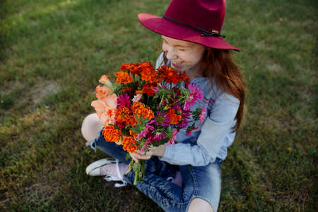 a beautiful teenage girl in a big red hat is sitting with a bouquet of different flowers on the green grassの写真素材