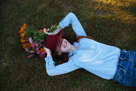 a beautiful teenage girl in a big red hat is lying on the grass with a multicolored bouquet of flowers. top viewの写真素材