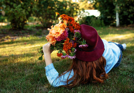 a beautiful teenage girl in a big red hat is lying on the grass with a multicolored bouquet of flowers. side viewの写真素材