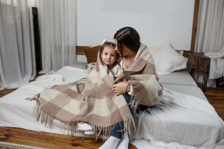 A happy young mother and her little daughter are sitting on the edge of the bed, covered with a blanket and smiling at the camera.の写真素材