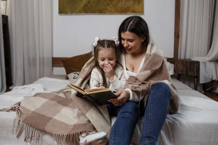 a loving family. A pretty young mother is reading a book to her daughter at home on the bed covered with a blanket.の写真素材