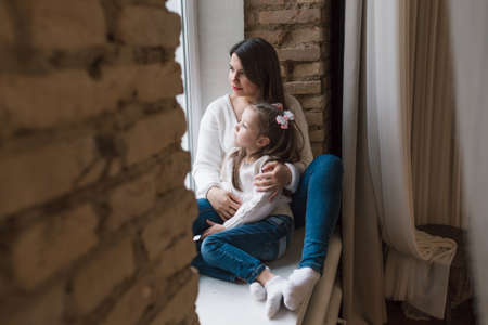 A young beautiful mother is sitting on a wooden windowsill in a white sweater and jeans with her arms around her young daughter.の写真素材