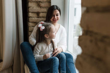 A young beautiful mother is sitting on a wooden windowsill in a white sweater and jeans with her arms around her young daughter.の写真素材