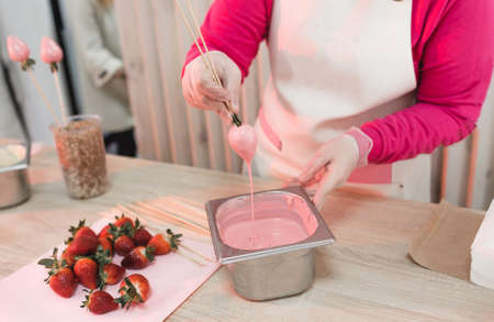 A pastry chef dips strawberries on a wooden skewer into a container with pink chocolate. Preparation of desserts.の写真素材