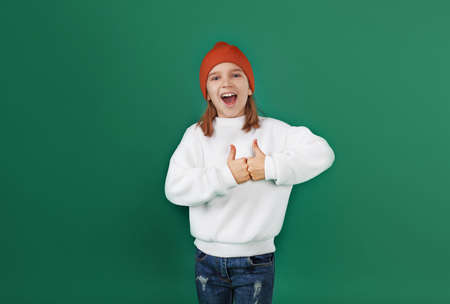 a little girl in a white sweater and a red hat laughs and shows her thumbs up on a green isolated background. children's emotionsの写真素材
