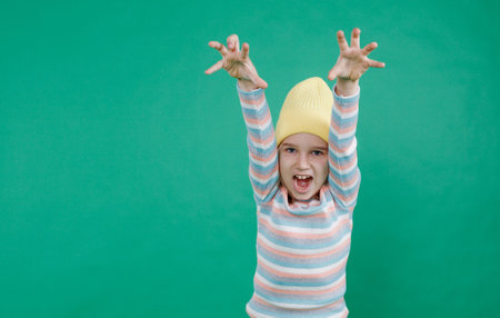 a little boy with long hair in a yellow hat shouts loudly and raises his hands up against an isolated green background.の写真素材