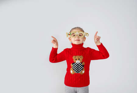 a little girl in a red sweater and funny Christmas glasses poses on a white background with her hands up.の写真素材