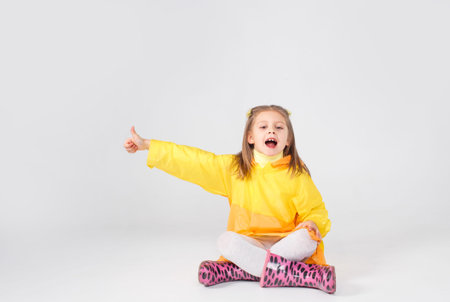 a little girl in a yellow raincoat and boots is sitting on a white background pointing her finger...