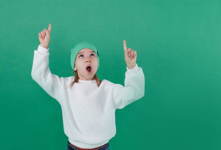 a joyful boy in a white sweater and hat stands with his hands up on a green background