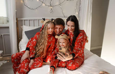 Father, mother and two little daughters in bed are photographed looking at the phone. Happy family in red Christmas pajamas in the bedroom on Christmas morning.の写真素材