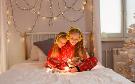 beautiful little girls sitting in bed in bright Christmas pajamas listening to music with headphones...