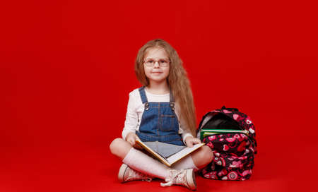 a schoolgirl girl with a backpack is sitting cross-legged on a red isolated background reading a bookの写真素材