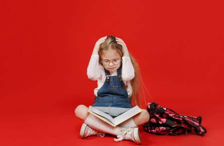 schoolgirl girl is sitting with a book upset with her hands on her head, worrying on a red...