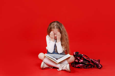 a little schoolgirl girl with a book is sitting on a red isolated background, covering her face with her hands.の写真素材