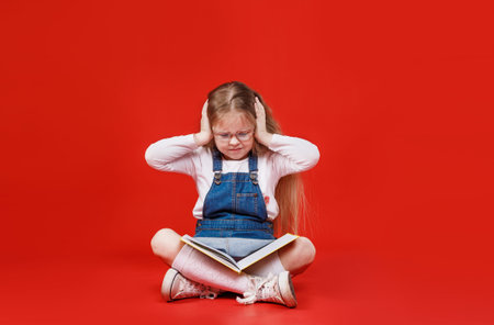 schoolgirl girl is sitting with a book upset with her hands on her head, worrying on a red...