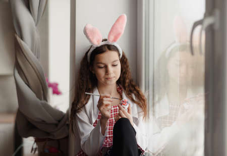 a beautiful girl with rabbit ears on her head draws eggs for an Easter basket on the windowsill against the windowの写真素材