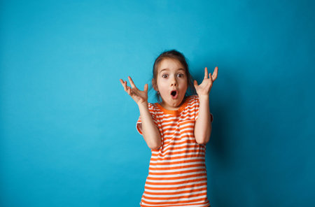Portrait of a surprised cute little girl standing isolated on a blue background. looking at the...