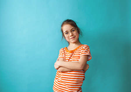a little beautiful girl with glasses stands with her arms crossed on a blue backgroundの写真素材