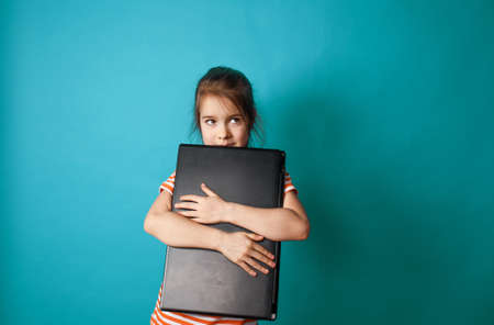 a little beautiful girl hugs a large laptop in her hands on a blue isolated backgroundの写真素材