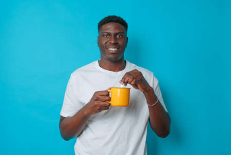 A young African American man drinks coffee from a yellow cup stirring with a spoon on a blue isolated background.の写真素材