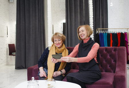 Two elderly women smile and take pictures of themselves on a smartphone while sitting on a sofa in a...