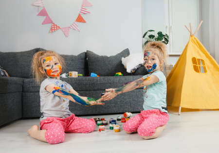 little twin girls got dirty with paints for drawing sitting at home on the floor in the room.の写真素材
