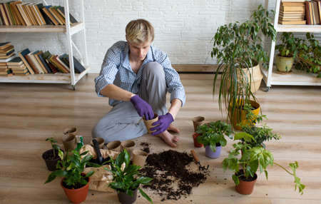 An attractive young man takes care of home plants, sits on the floor in the living room, transplants flowers, pours earth into flower pots. The concept of gardening at home.の写真素材