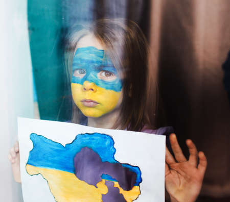 a little girl with a painted Ukrainian flag on her face is standing behind the glass with sad eyes holding a burnt map of Ukraine in her hands. Children against warの写真素材