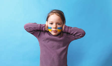 a little frightened girl with a Ukrainian flag painted on her cheek covers her ears with her hands so as not to hear anything on an isolated blue background. Stop the war in Ukraineの写真素材