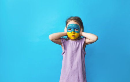 a little frightened girl with a Ukrainian flag painted on her face covers her ears with her hands...